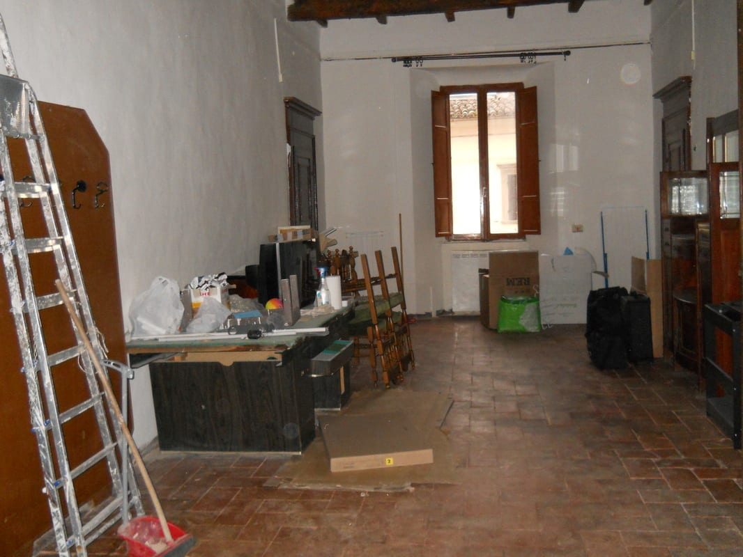 A large bare hall with tiled floor, stone window surrounds, and assorted old furniture and boxes piled along the walls before renovation.