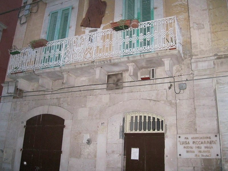 Exterior of a two-storey stone building with ornate iron balcony, green shutters, and a commemorative plaque beside the entrance.