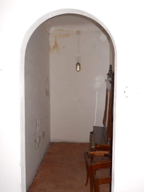 A plain whitewashed corridor ending in an arched doorway, with a bare hanging bulb and a wooden chair to the right.