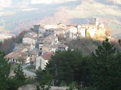 Hilltop Italian village of stone buildings and a church tower seen from across a wooded valley.