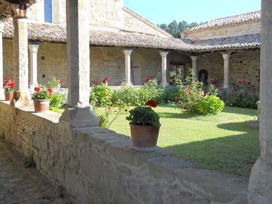 A sunlit cloister garden with potted red geraniums along a stone arcade and a lawn planted with flowering shrubs.