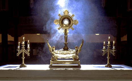 A gold monstrance on a white altar flanked by two candelabra, with incense smoke rising and diffuse blue light in the dark background.