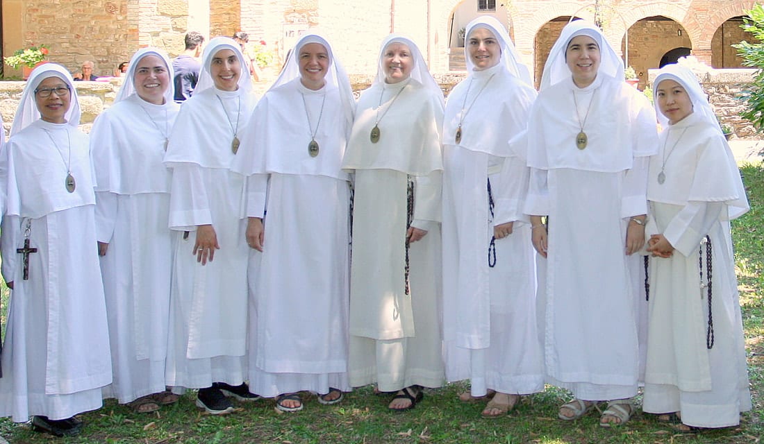Eight sisters in white habits and veils posing together in a sunlit cloister courtyard.