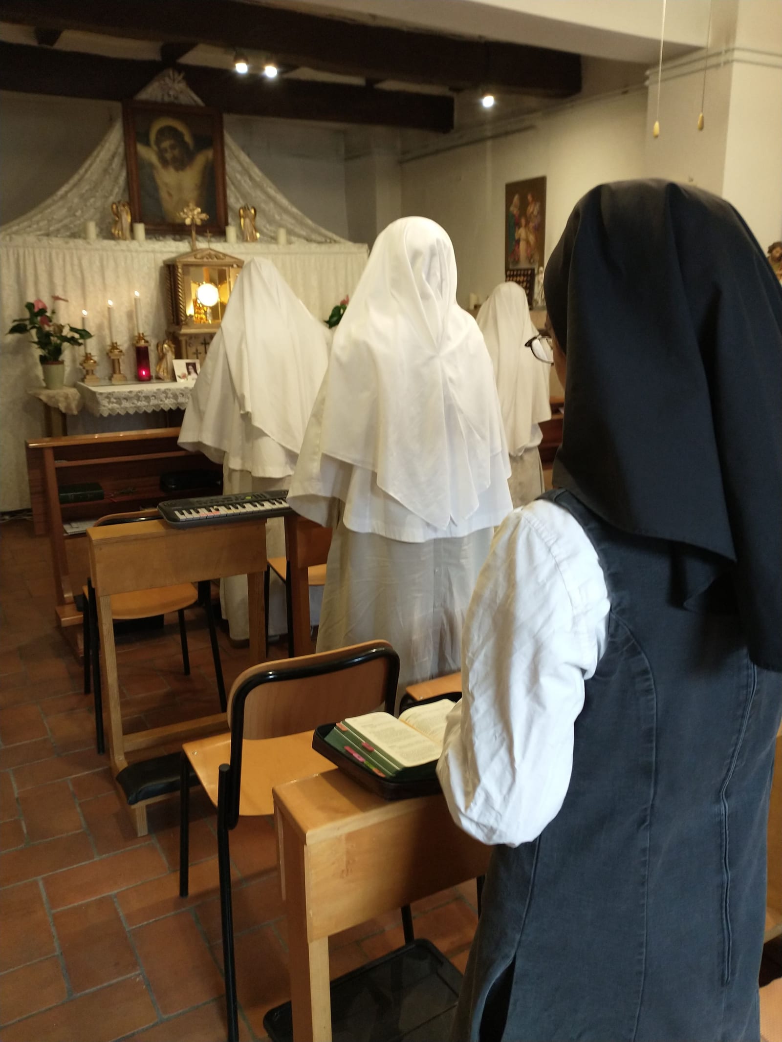 Suore dai lunghi veli bianchi in piedi davanti al Santissimo Sacramento esposto sull'altare, e una postulante dal velo nero che segue le preghiere sul suo libro.