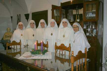 Six sisters in white habits standing together at a table with a lit Advent wreath, inside a room with wooden furnishings and a religious artwork visible in the background.