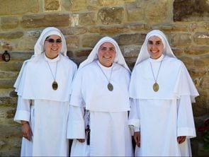 Three sisters in white habits and veils standing together outdoors against a stone wall, smiling.