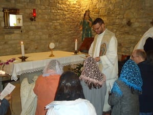 A priest distributing communion to kneeling figures in a small stone chapel with an altar and statue visible behind him.