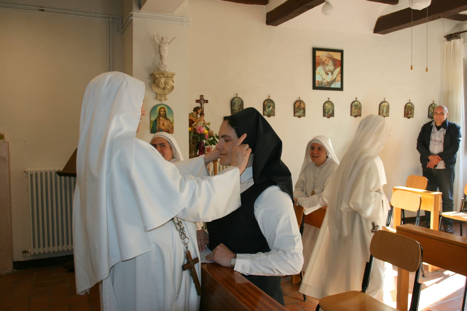 Una suora in bianco pone il velo nero su una postulante a occhi chiusi, mentre altre suore guardano sorridendo nella cappella.