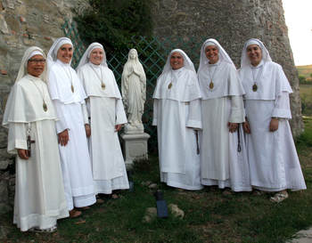 Six sisters in white habits gathered outdoors beside a stone wall and a small Marian statue, smiling for a group portrait.