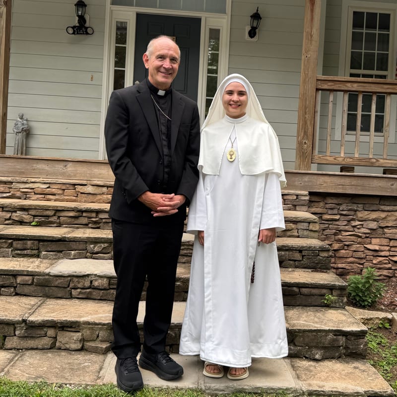 Sister Faustina in the white habit on the day of her first vows, standing beside a priest on the steps of a house with a porch.
