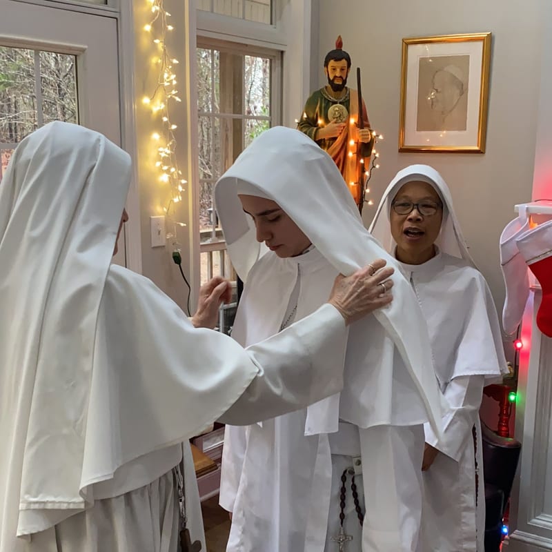 Sisters in the white habit settle the veil of a newly clothed novice, in a room with a statue of a saint and lights strung along the window.