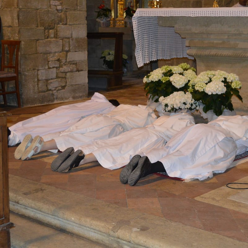 Sisters in white lying prostrate on the floor before the altar during their perpetual profession, with white hydrangeas beside them.