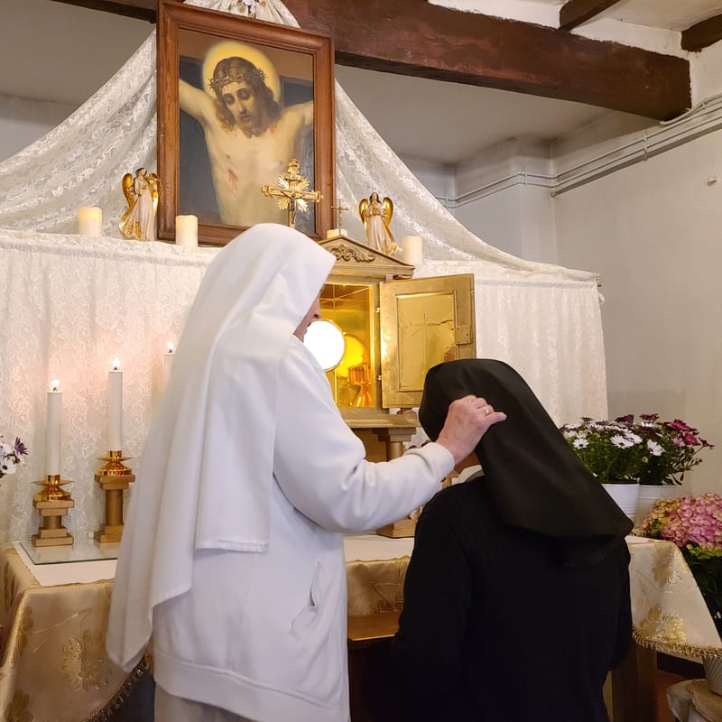A sister in the white habit places a black veil on a kneeling postulant before an altar, beneath a painting of Christ crowned with thorns.