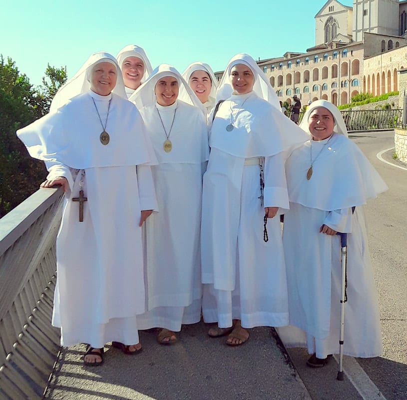 Six sisters in white habits posing on a bridge or terrace outside a large stone basilica on a clear day.