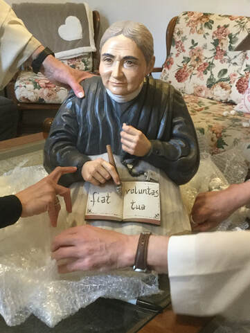 A painted or molded figurine of a seated elderly woman writing in an open book inscribed ‘Fiat voluntas tua,’ with several hands reaching toward her.