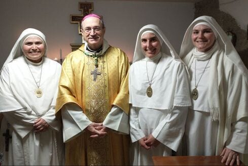 A bishop in gold vestments standing with three sisters in white habits inside a stone room.