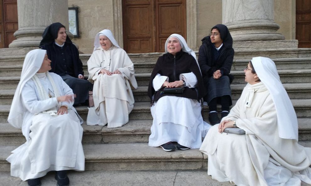 A group of sisters and lay visitors in black and white habits seated and standing on the broad stone steps of a large church doorway, talking and laughing.