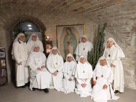 A community of nine sisters in white habits posed in two rows inside a stone-walled room, with an image of Our Lady of Guadalupe on the wall behind them.