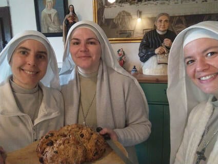 Three sisters in light grey-white habits holding a loaf of bread and smiling in a kitchen, with a framed portrait visible on the wall behind them.