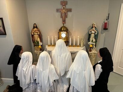 Sisters in white veils kneeling in prayer before a small altar with candles, a monstrance, and statues of the Sacred and Immaculate Hearts under a crucifix.