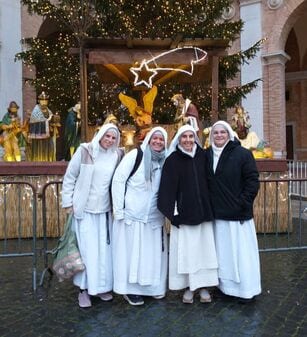 Four sisters in white habits standing together in front of a large outdoor nativity scene with a lit Christmas tree at night.