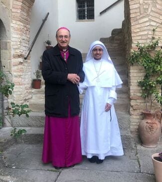 A bishop in purple cassock and a sister in a white habit standing together in a stone courtyard.