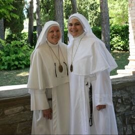 Two sisters in white habits standing together outdoors among trees and a stone balustrade, smiling.
