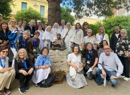 A large group of pilgrims and sisters gathered outdoors around a bronze seated statue of the Servant of God Luisa Piccarreta.