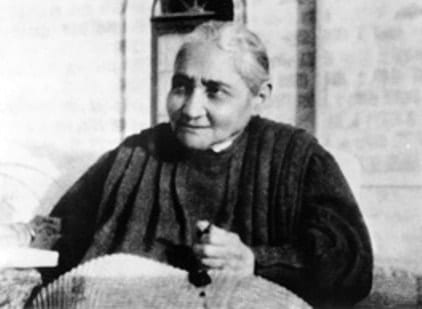 Black-and-white photograph of the Servant of God Luisa Piccarreta seated outdoors, looking to one side, with lace or needlework resting in her lap.