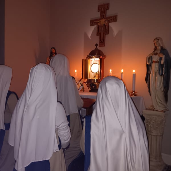 Sisters kneel in adoration before the Blessed Sacrament exposed in a monstrance, lit by candles, beside a statue of Our Lady.
