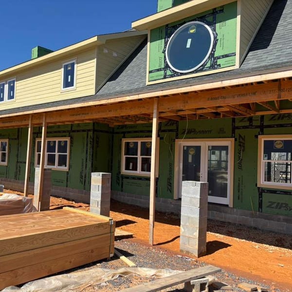 The new monastery in Maryville under construction: a long covered porch and a gable with a round window, with building materials stacked in front.
