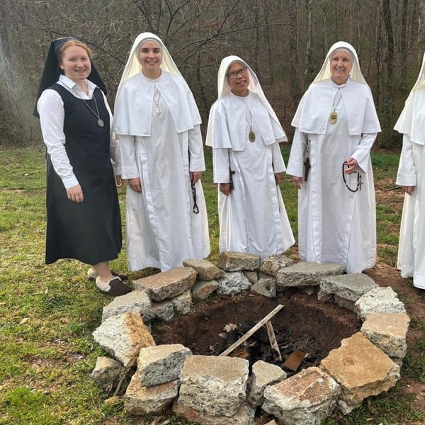 Four sisters in white and one in black stand smiling around a stone fire pit in the woods.