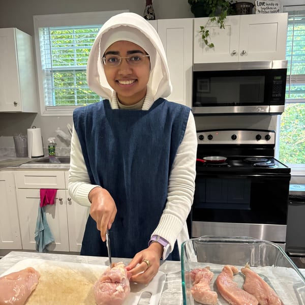In the kitchen, a smiling young woman in a white veil and blue jumper prepares chicken.