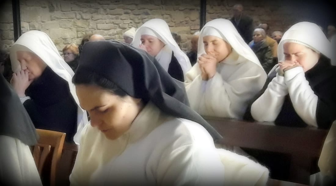 Several sisters in white and dark habits bowed in prayer at wooden pews inside a stone chapel.