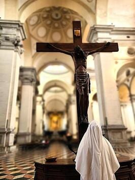 A sister in a white habit kneeling before a large wooden crucifix inside a bright church nave.