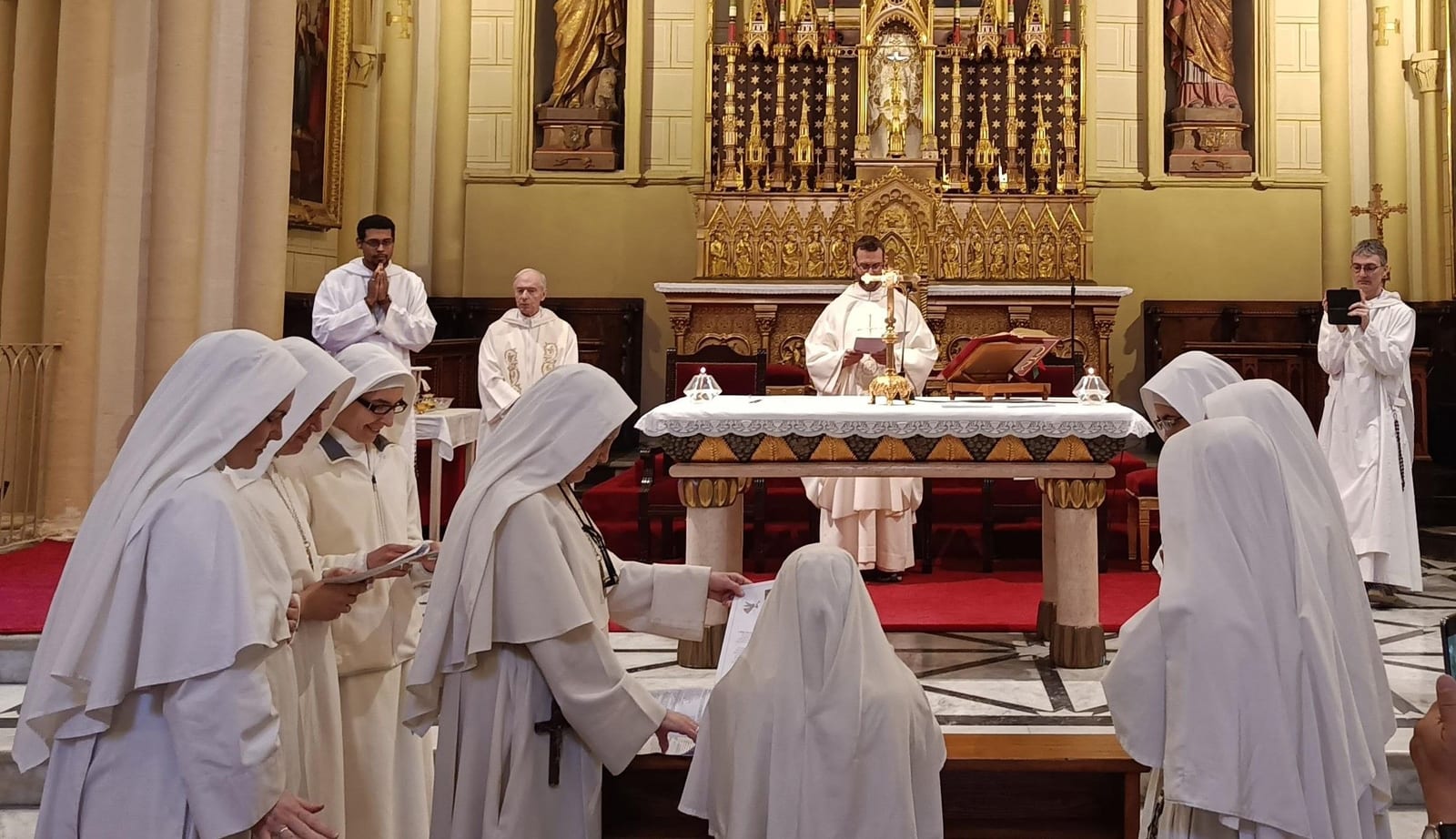 In una chiesa con un altare maggiore dorato, una suora porge un foglio a un'altra inginocchiata davanti all'altare durante una professione dei voti, mentre un sacerdote, alcuni monaci e le suore guardano.