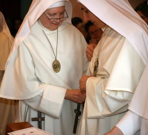 Two sisters in white habits, one presenting a cross to the other during a ceremony.