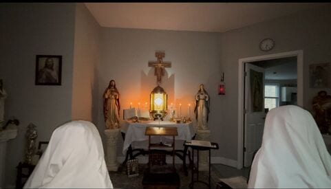 Two sisters in white veils kneeling in prayer facing a small illuminated altar with a monstrance and flanking statues in a dimly lit room.